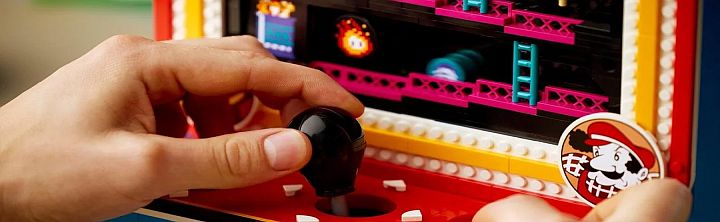 Person's hand on a black joystick in a colorful Lego arcade cabinet with a Mario sticker nearby